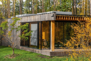 Image of a modern modular home with wooden exterior and large glass windows, surrounded by grassy area and autumn trees, featuring a cozy interior with warm lighting and a chair.