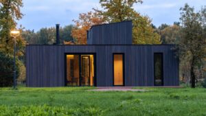 Image of a modern modular home with dark wood exterior, large windows, and warm interior lighting, set in a grassy area with autumn trees and a streetlamp, showcasing its sleek design.