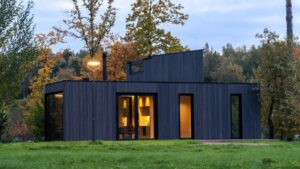 Image of a modern modular house with dark wood exterior and large glass doors, illuminated warmly inside, set in a grassy area with autumn trees.