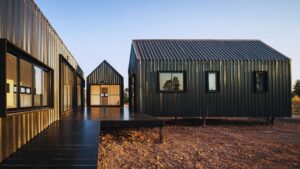 Modern black corrugated metal tiny house cluster with pitched roofs, elevated on stilts, connected by wooden deck walkway at sunset.