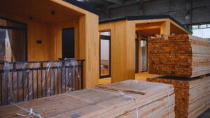 Stacks of lumber and building materials in front of partially completed wooden modular home units inside a factory warehouse.