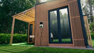 Modern wooden modular tiny home with EV charger plugged in, surrounded by greenery and pergola. Sustainable off-grid living.