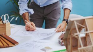 Architect customizing modular home design: hands sketching detailed blueprints with pencil, surrounded by small-scale house model on workspace table.